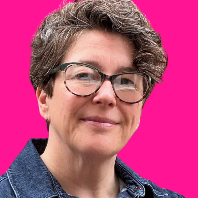 headshot of a middle-aged white woman with long brown hair and glasses and wearing a blu denim shirt. Her arms are folded and she's smiling.