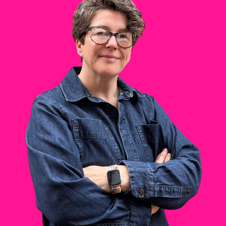 headshot of a middle-aged white woman with long brown hair and glasses and wearing a blu denim shirt. Her arms are folded and she's smiling.