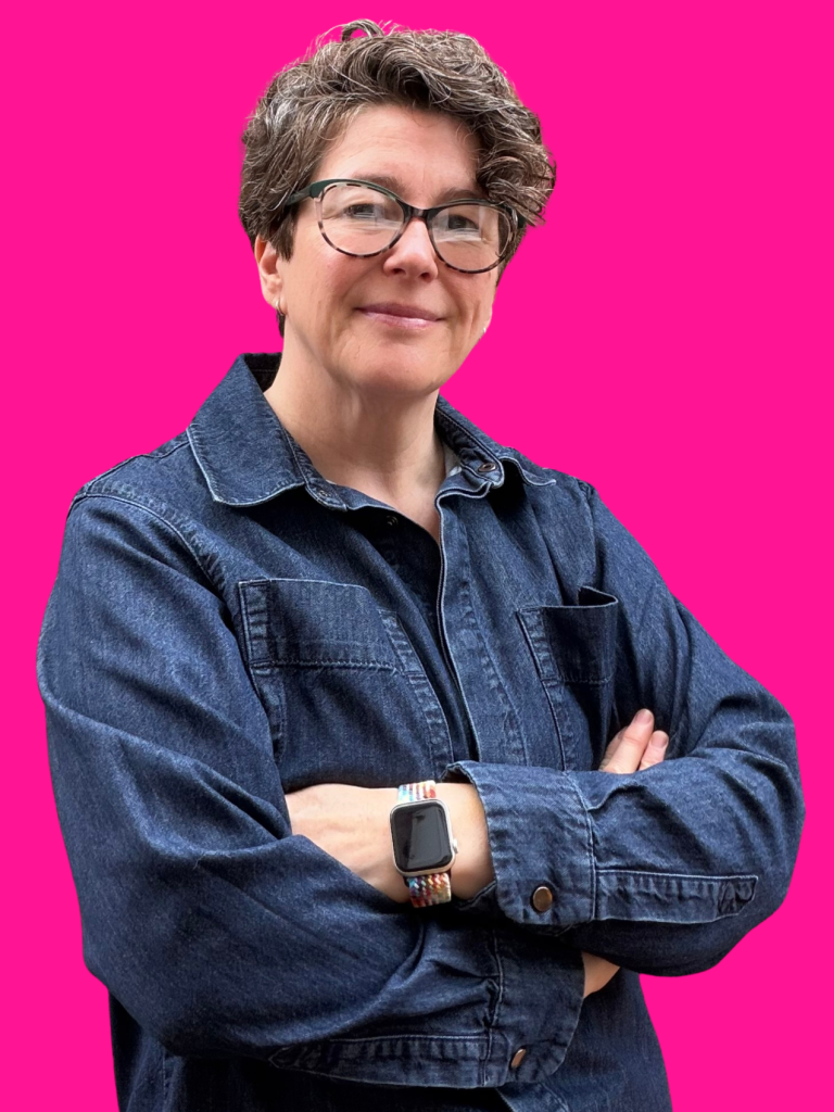 headshot of a middle-aged white woman with long brown hair and glasses and wearing a blu denim shirt. Her arms are folded and she's smiling.