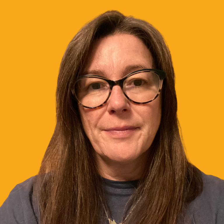 headshot of a middle-aged white woman with long brown hair and glasses and wearing a blu denim shirt. Her arms are folded and she's smiling.