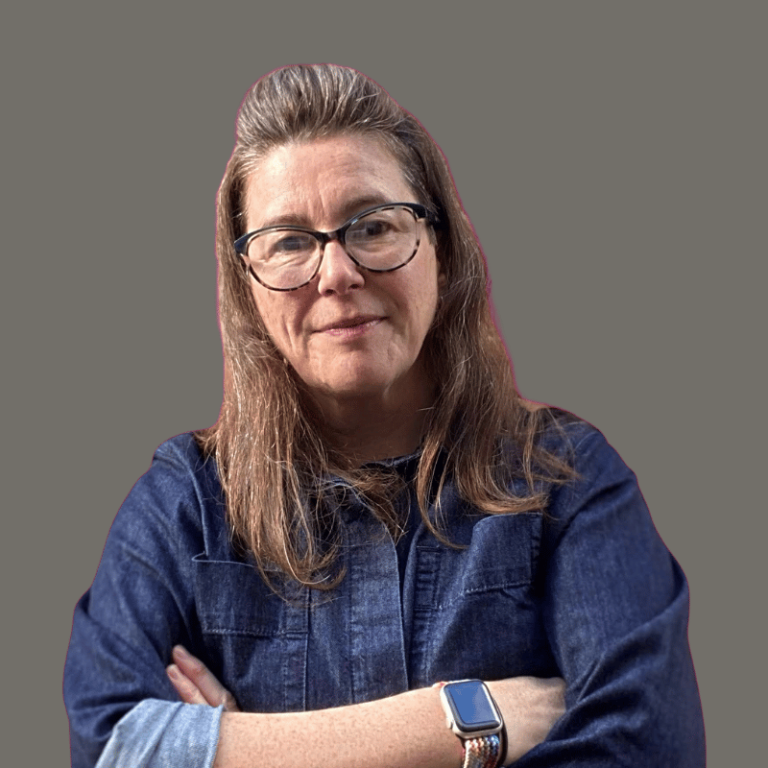headshot of a middle-aged white woman with long brown hair and glasses and wearing a blu denim shirt. Her arms are folded and she's smiling.